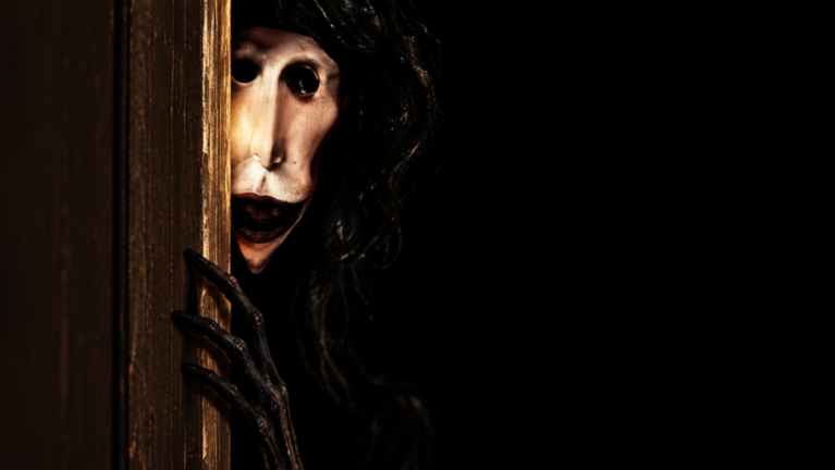 A pale-faced creature with long black hair peeking through a wooden door, gripping the edge with long dark fingers.